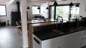 A modern kitchen with a central island, black pendant lights, and a view of the outdoors through large windows.
