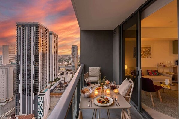 Un balcón con una mesa para dos personas, con vistas a un paisaje urbano al atardecer.