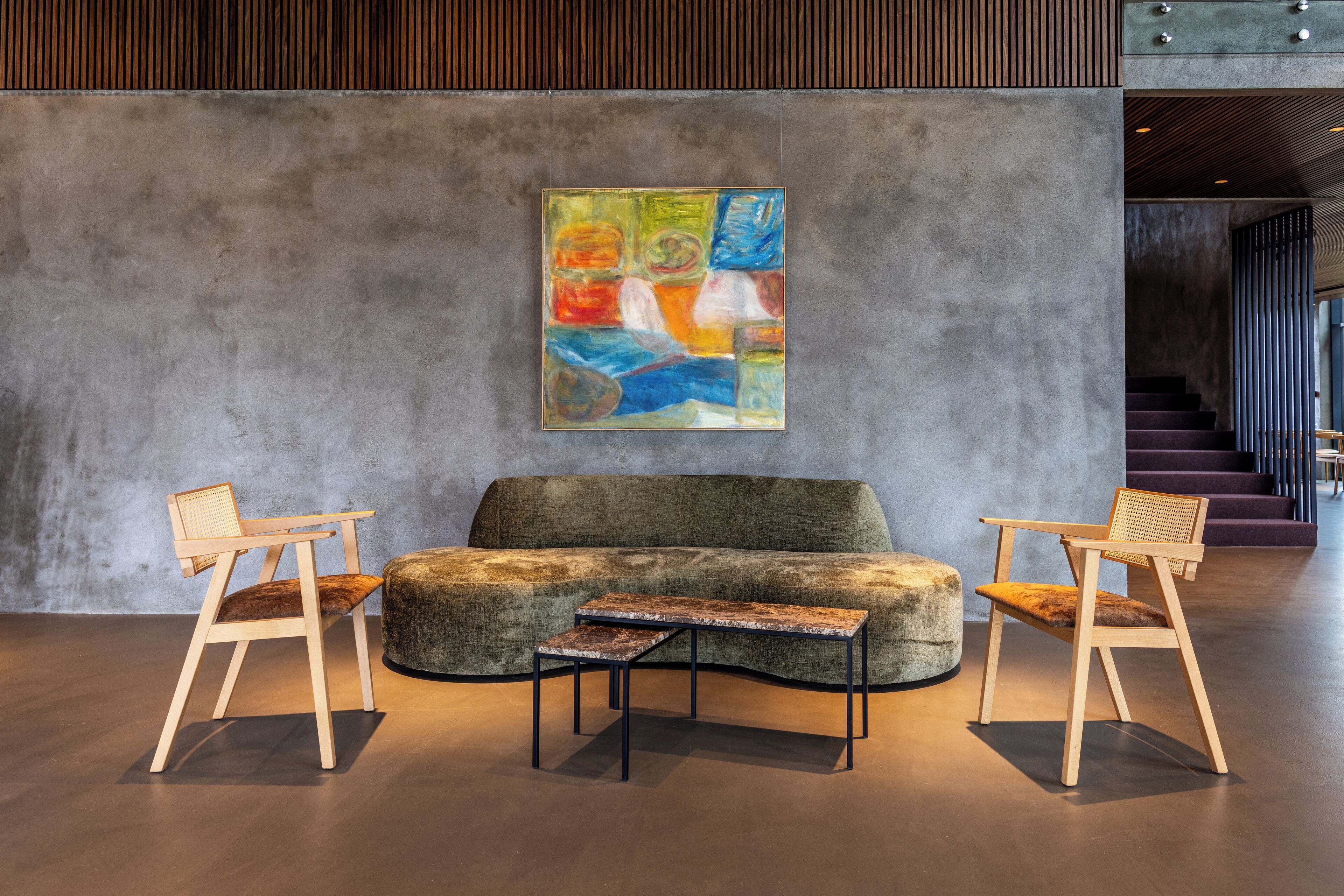 Een moderne lobby met een grijze bank, twee houten stoelen, een klein tafeltje en een kleurrijk abstract schilderij aan de muur.