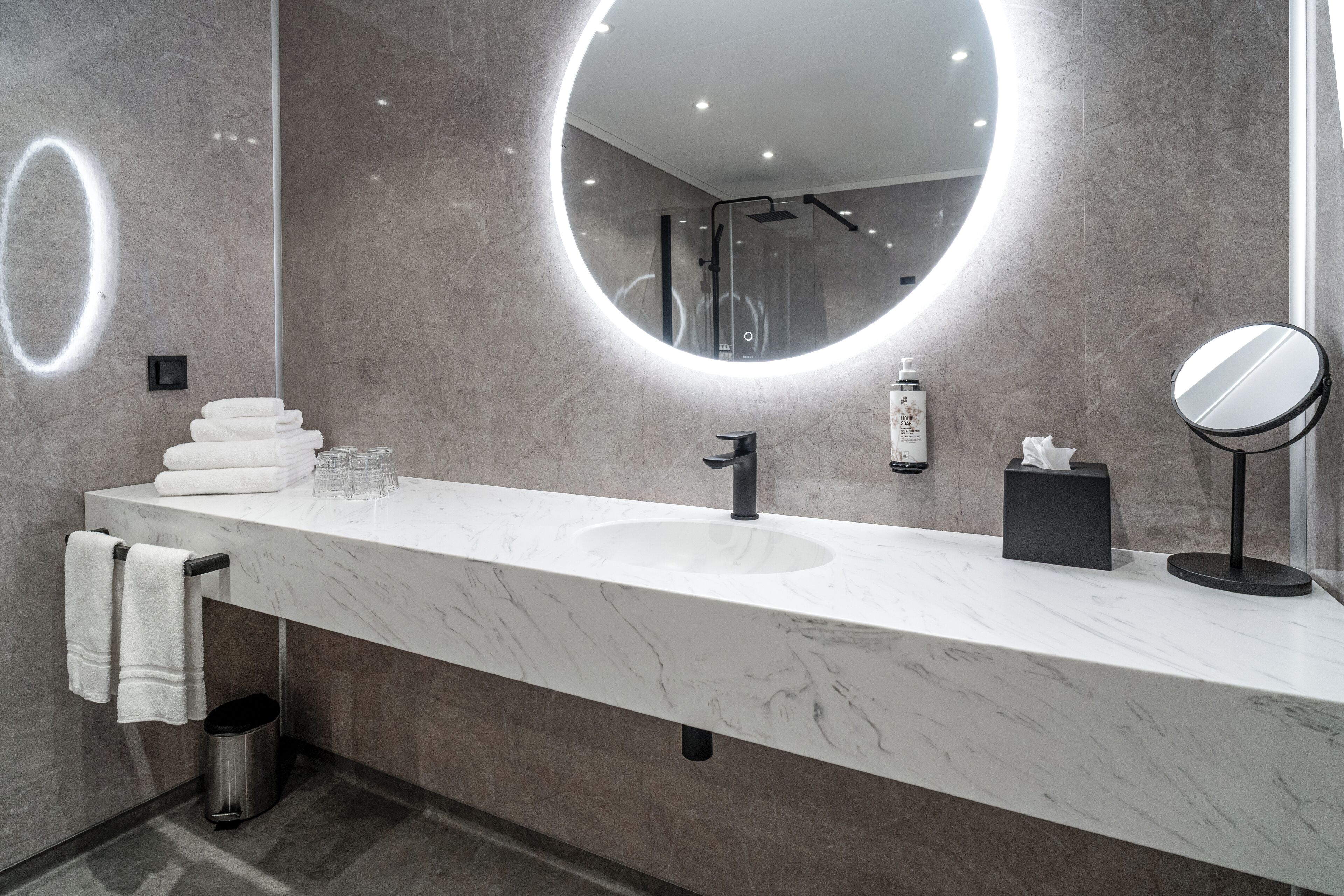 A modern bathroom with a marble countertop, a round mirror, a handheld showerhead, and a towel rack.