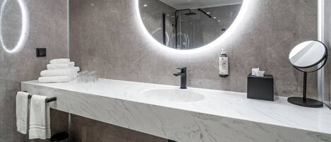 A modern bathroom with a marble countertop, a round mirror, a handheld showerhead, and a towel rack.