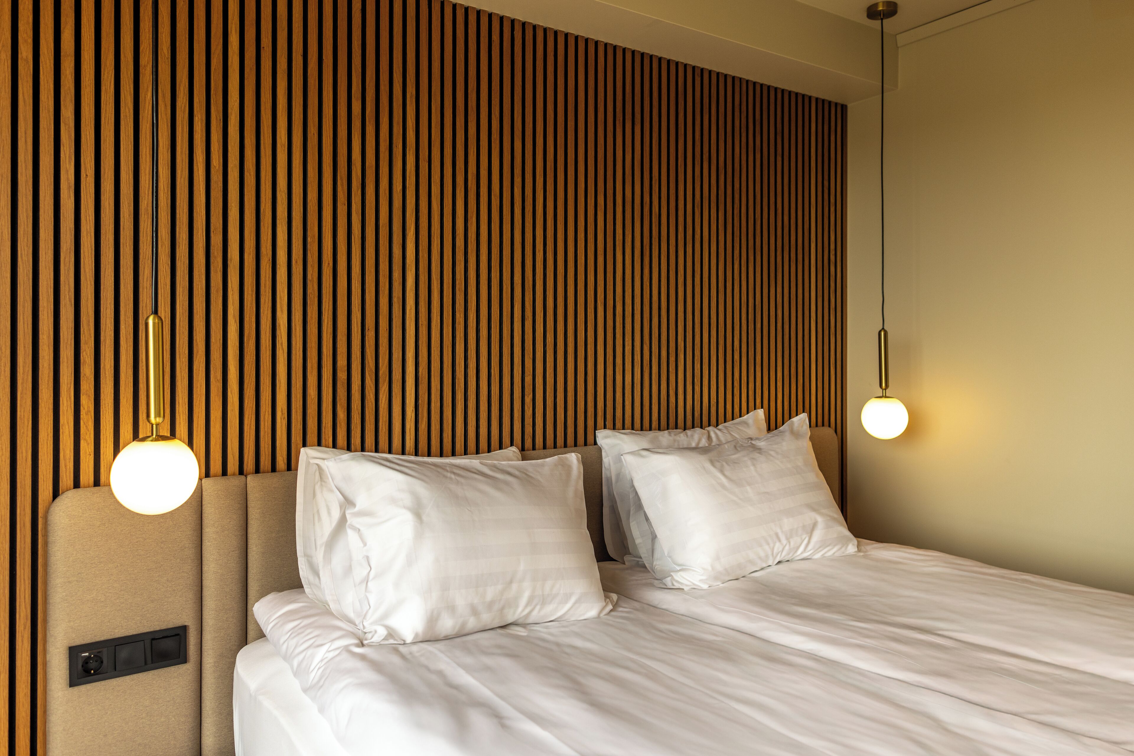 A neatly made bed with white linens and two pillows, a wooden slat headboard, and two pendant lights.