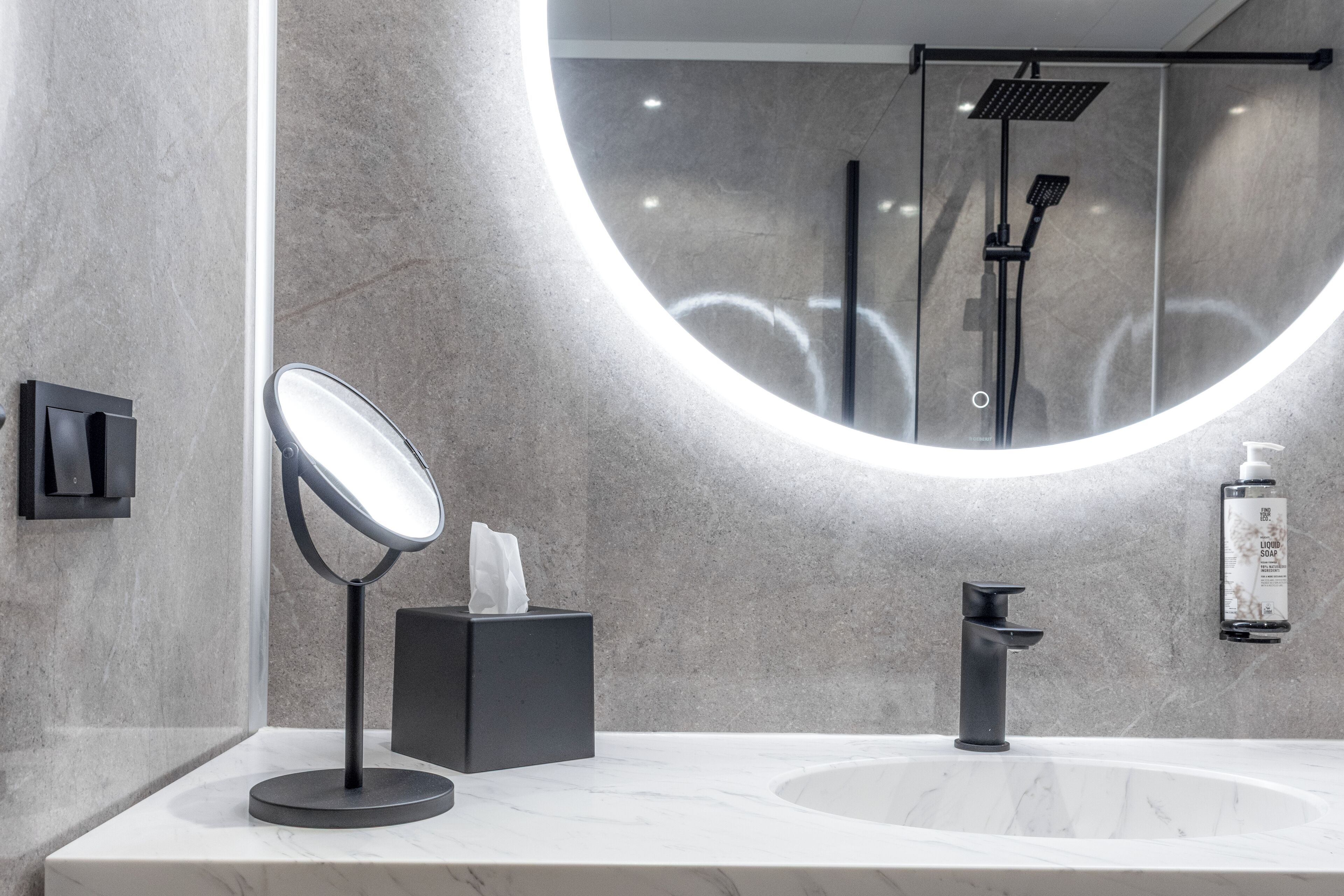 A modern bathroom with a round mirror, a black soap dispenser, and a handheld showerhead.