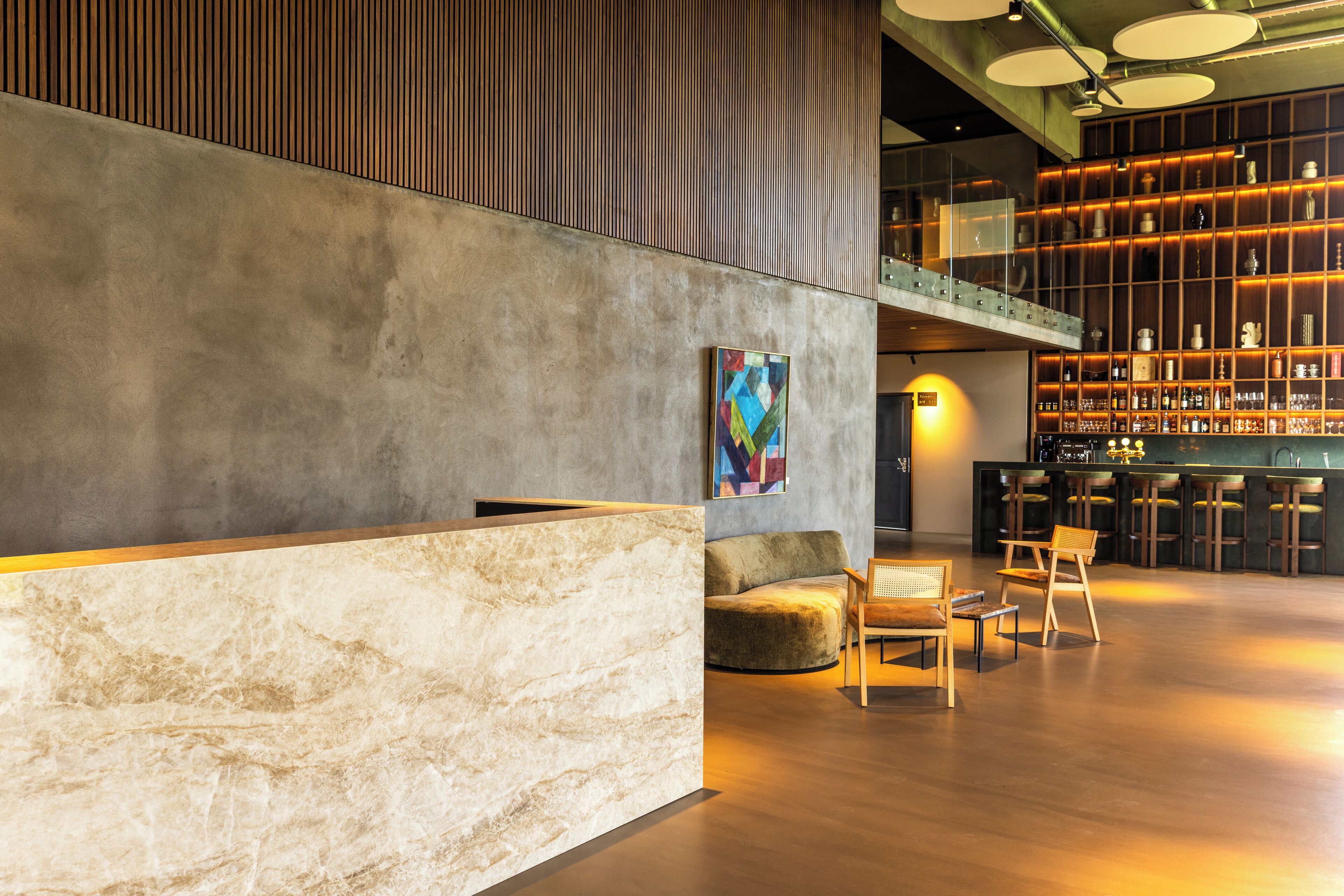 Un vestíbulo moderno con un mostrador de recepción de mármol, un cuadro abstracto colorido, una zona de estar con sillas de madera y un bar con taburetes.