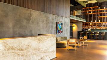 Un vestíbulo moderno con un mostrador de recepción de mármol, un cuadro abstracto colorido, una zona de estar con sillas de madera y un bar con taburetes.