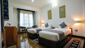 A hotel room with two beds, a desk, a chair, and a large window with curtains. - Hotel Lhading (Thimphu)