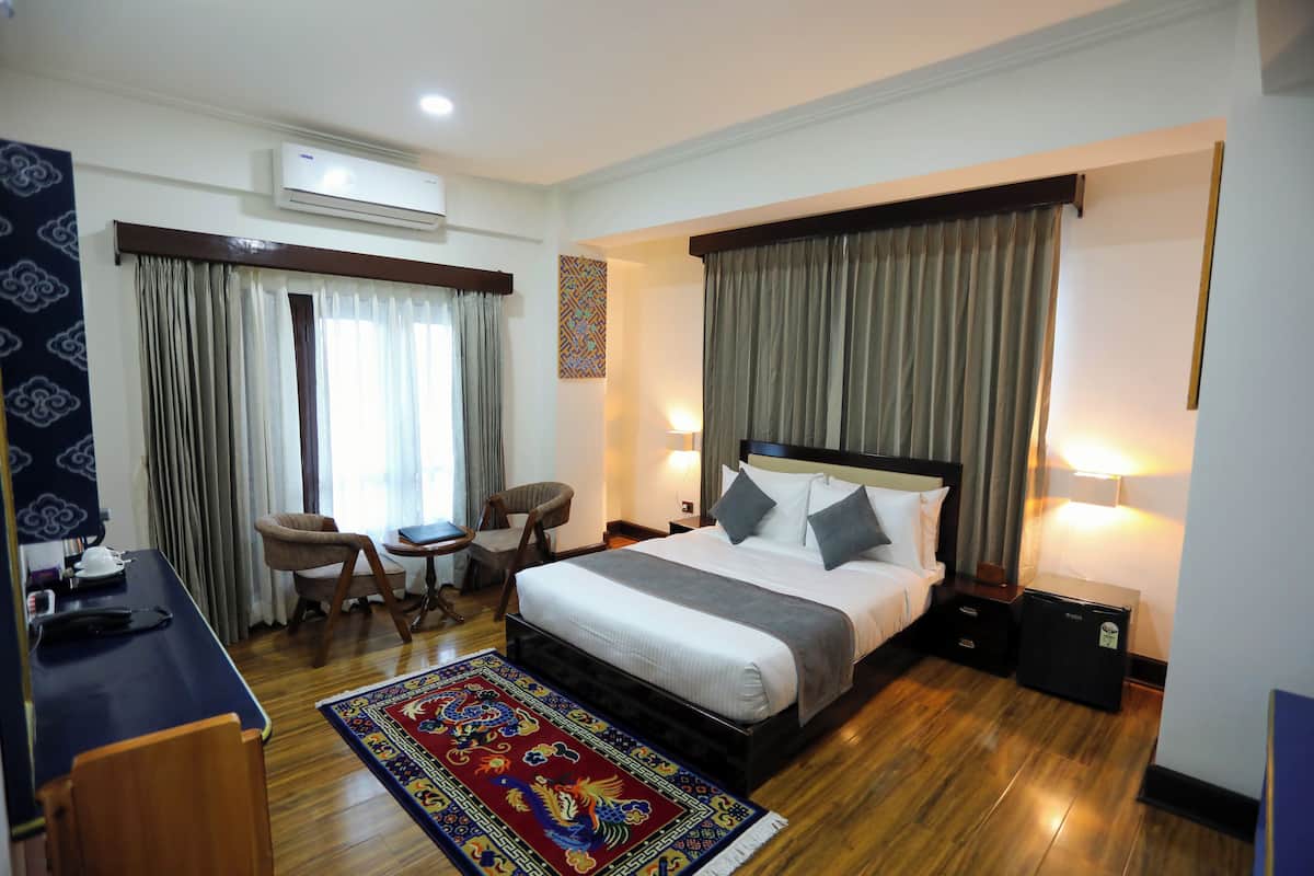 A hotel room with a bed, bedside tables, a desk, chairs, a rug, and an air conditioning unit.