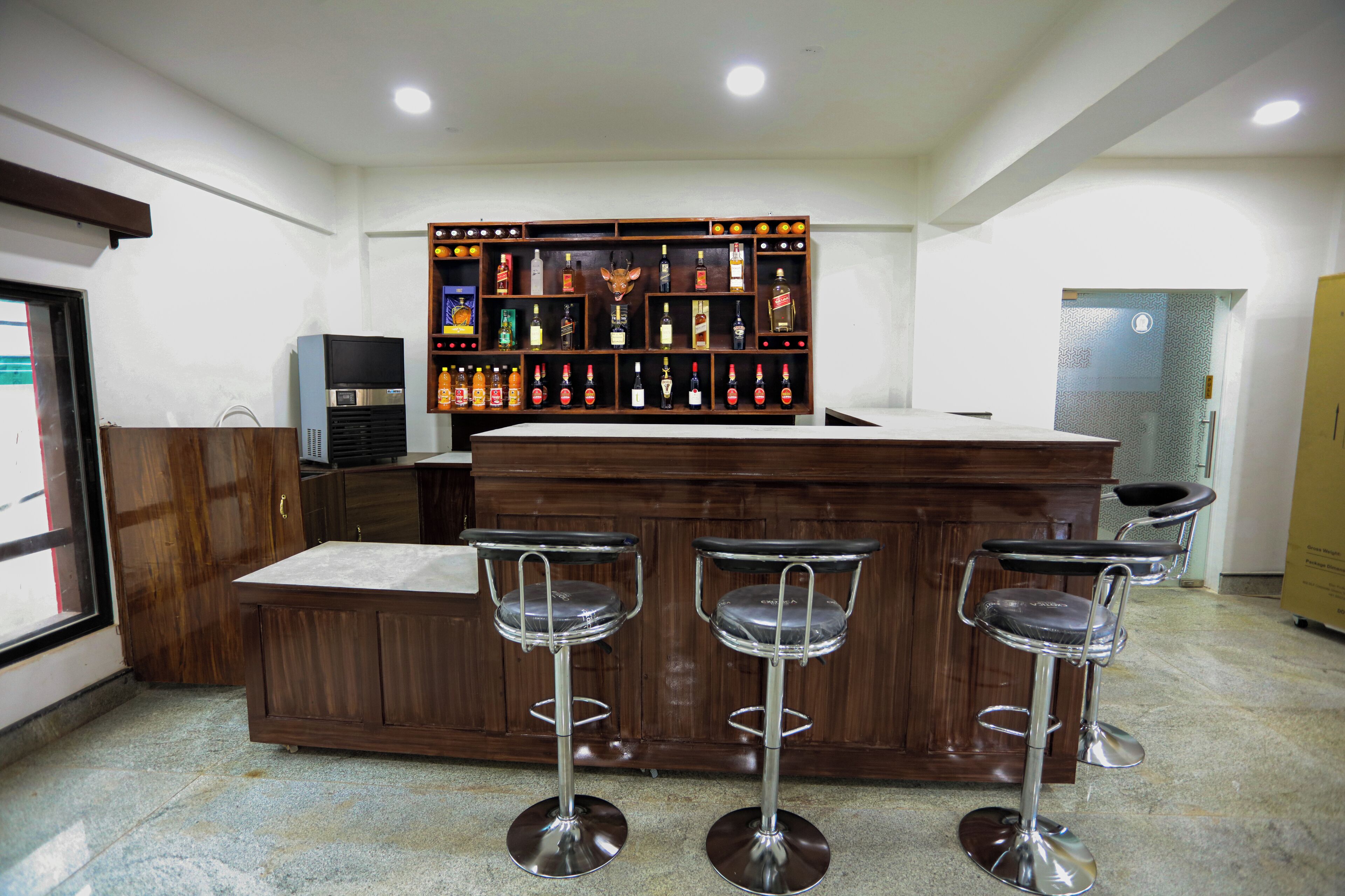 Una zona de bar con mostrador de madera y tres taburetes. Se observa una estantería con varias botellas detrás del mostrador.