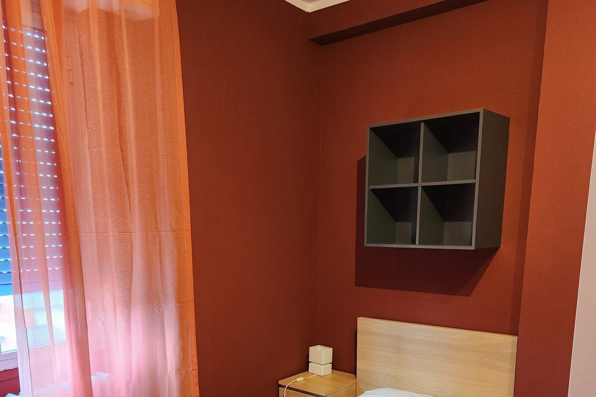 A single-bed room with a wooden headboard, a small bedside table, a wall-mounted shelf, and a radiator.