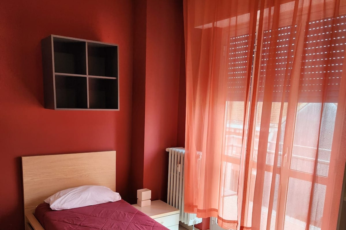 A bedroom with a wooden bed, red walls, a window with orange curtains, and a radiator.