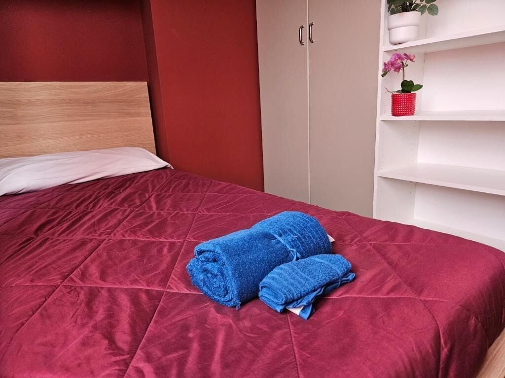 Una cama bien hecha con una colcha roja y dos toallas azules enrolladas.