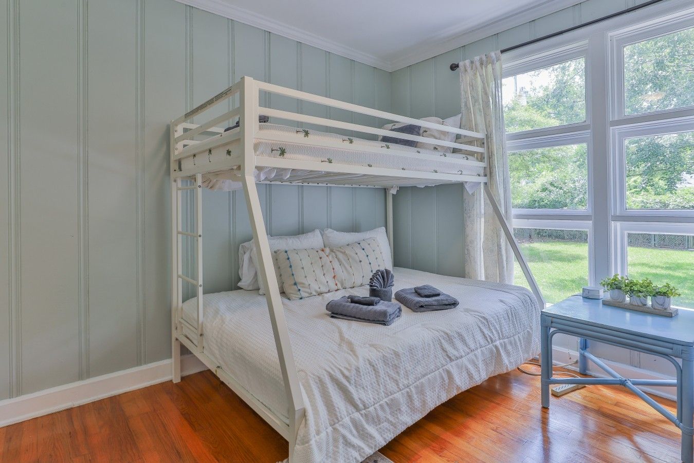 A bunk bed with a ladder, a bedside table with a plant, and a window with a view of greenery.