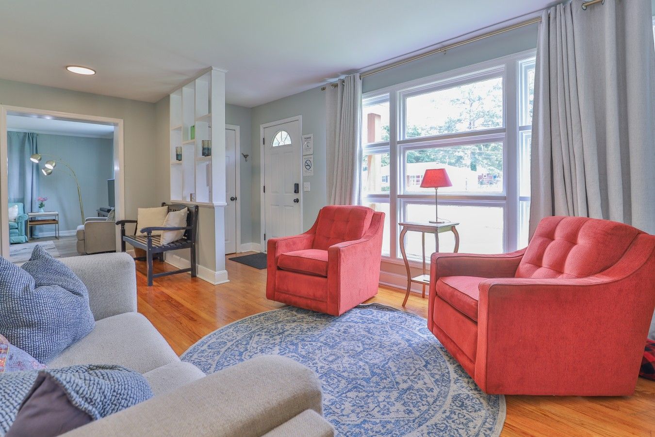 A living room with two red armchairs, a grey sofa, and a patterned rug. There is a window with curtains, a lamp on a side table, and a door with a sign.