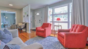 A living room with two red armchairs, a grey sofa, and a patterned rug. There is a window with curtains, a lamp on a side table, and a door with a sign.
