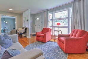 Una sala de estar con dos sillones rojos, un sofá gris y un tapete estampado. Hay una ventana con cortinas, una lámpara en una mesita auxiliar y una puerta con un cartel.