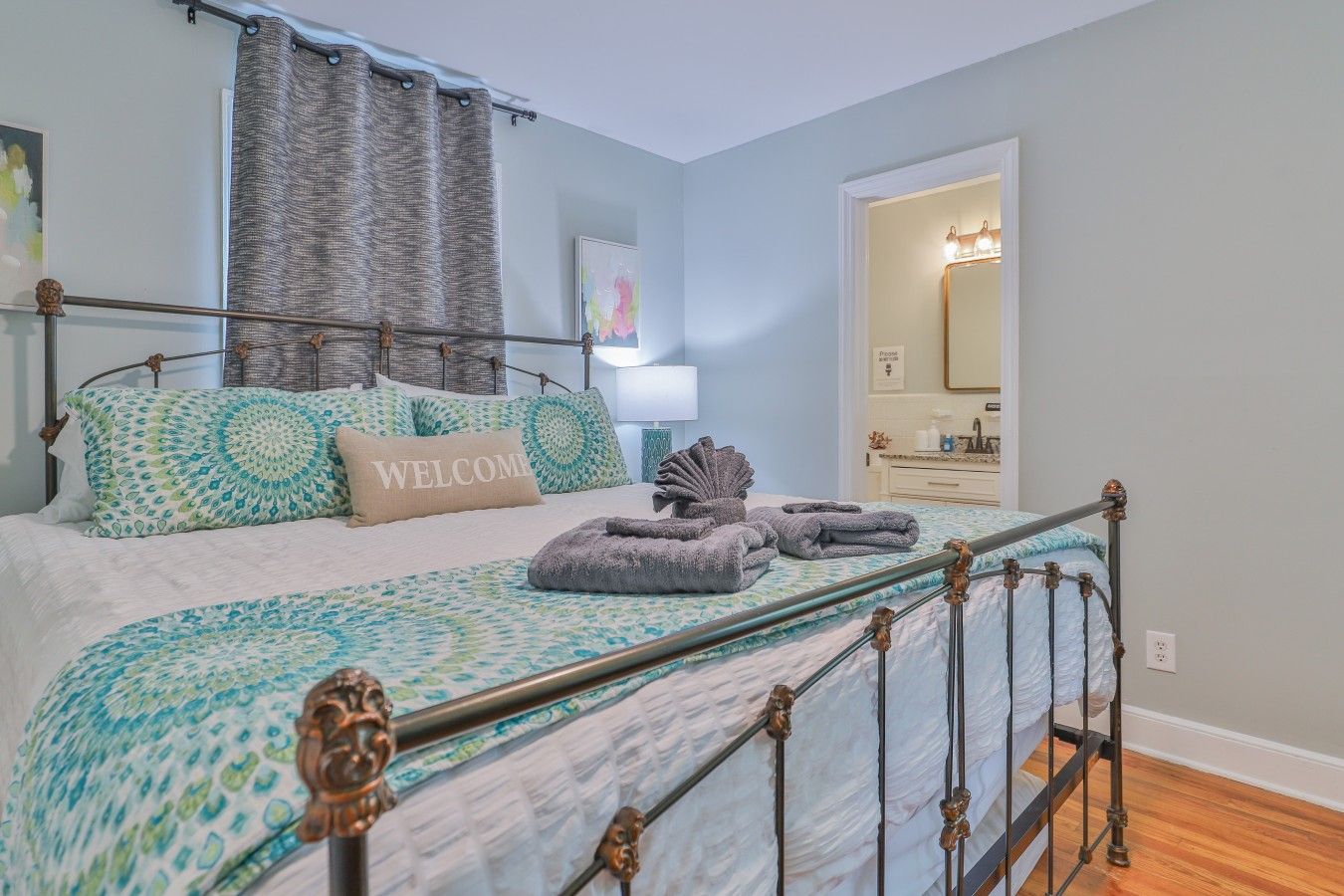 A bedroom with a metal bed frame, a bed with patterned bedding, a 'WELCOME' pillow, and a bathroom visible through an open door.