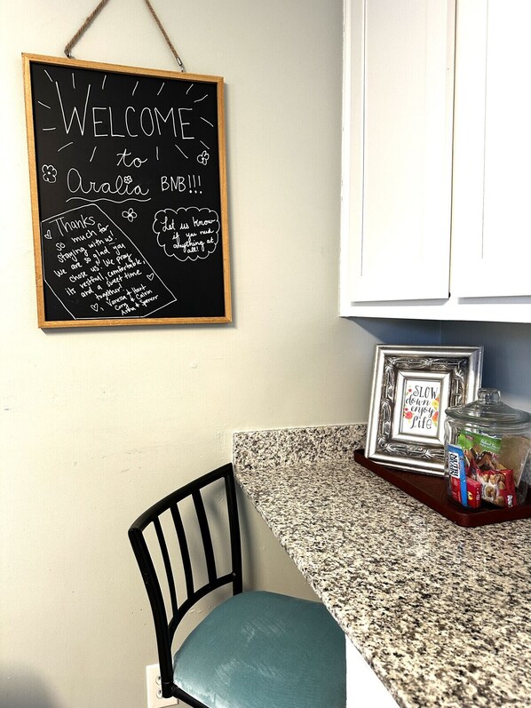 Una cocina con un letrero de pizarra que dice "WELCOME to Aralia BBB", una silla, una bandeja con aperitivos y una cita enmarcada sobre la encimera.