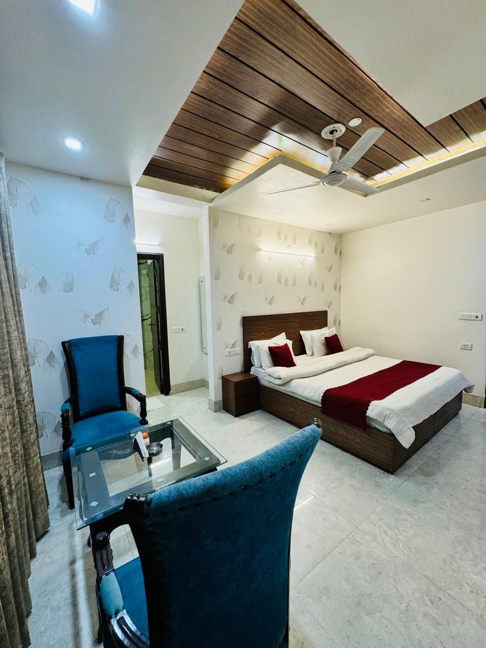 A hotel room with a bed, a chair, a glass table, and a ceiling fan.