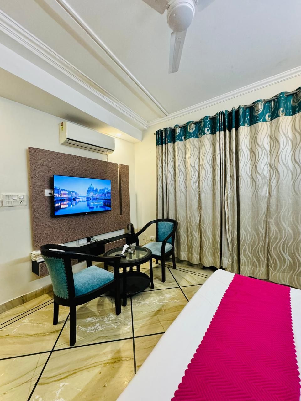 A hotel room with a TV mounted on the wall, a small table with chairs, a bed with a pink bedspread, and a window with patterned curtains.
