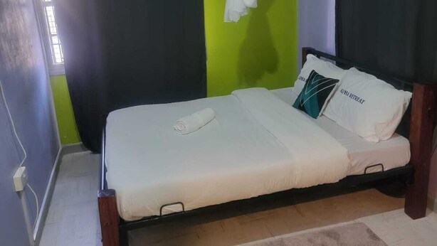 A single bed with a white mattress and a wooden frame, a brown rug, and a pair of green flip-flops on the floor. - ALPHA RETREAT (Kisumu)