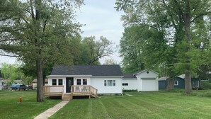 A single-story mobile home with a front porch and a small deck, surrounded by a grassy yard and trees.