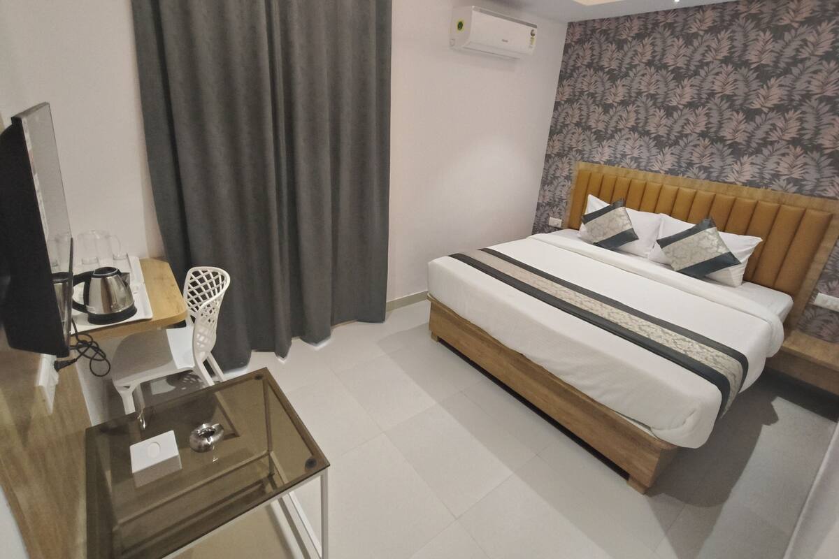 A modern hotel room with a bed, a desk with a coffee maker, a TV, and an air conditioning unit.
