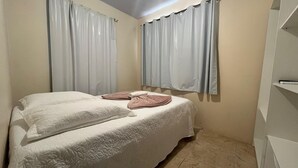 A small, single-bed room with a plain bedspread, two sets of towels, and light curtains on the windows. - AP 10 Ala Conceição Residencial Solavir (Bombinhas)