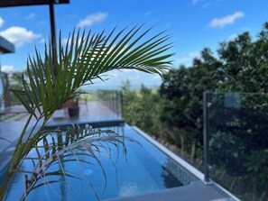Una piscina con ringhiera in vetro, circondata da una vegetazione rigogliosa e un cielo azzurro intenso.