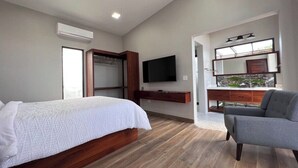 A modern bedroom with a bed, a gray armchair, a TV, and a wooden floor. - Villa 15 at Chial Reserve Near San Ignacio (San Ignacio)