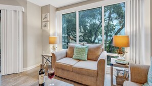Una sala de estar acogedora con un sofá color beige, una mesa de centro con una botella de vino y una copa, y vistas a árboles a través de amplias ventanas.