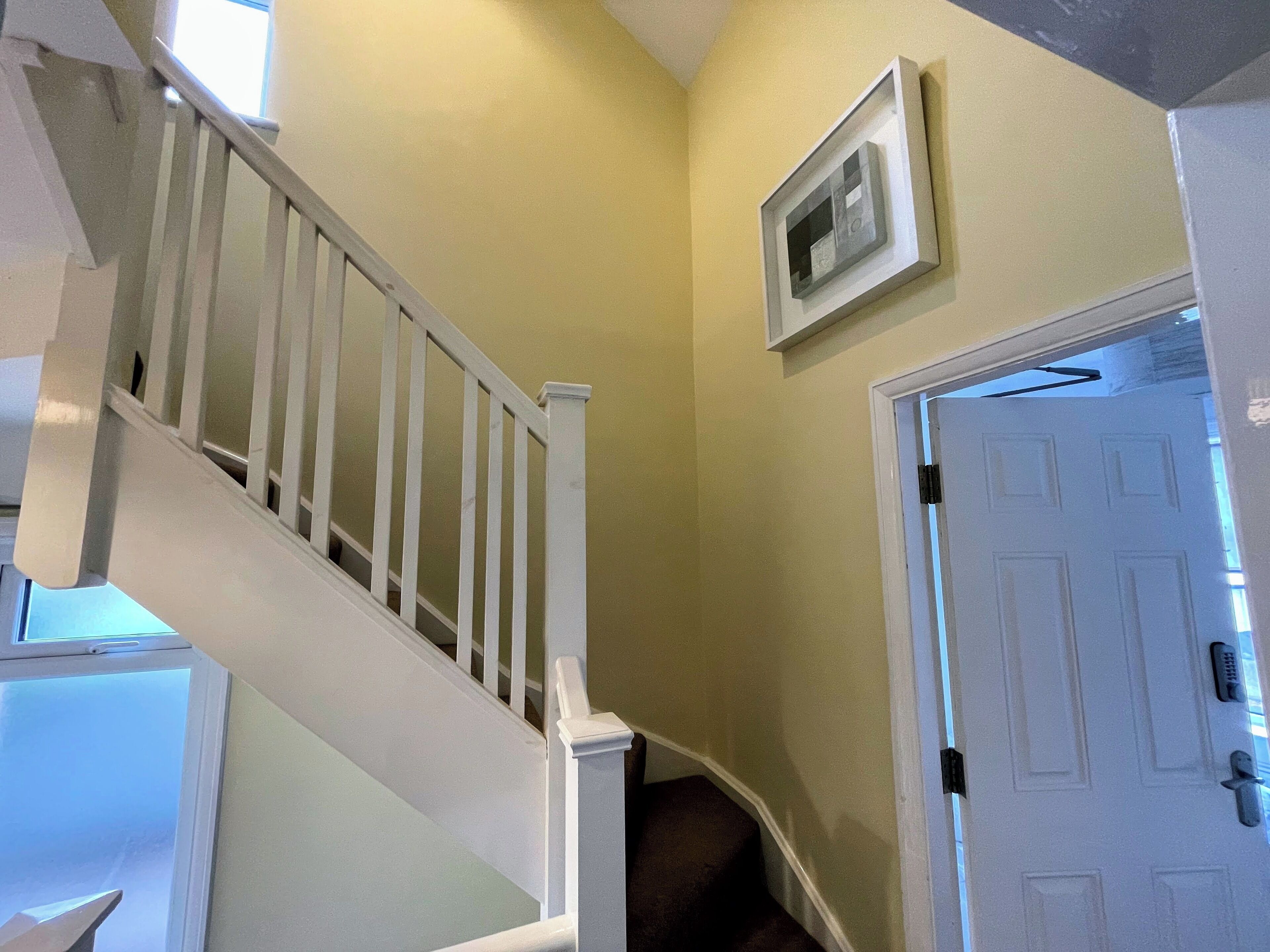 A staircase with white railings leading to an upper floor. A white door is visible to the right, and a framed picture hangs on the wall above the door.
