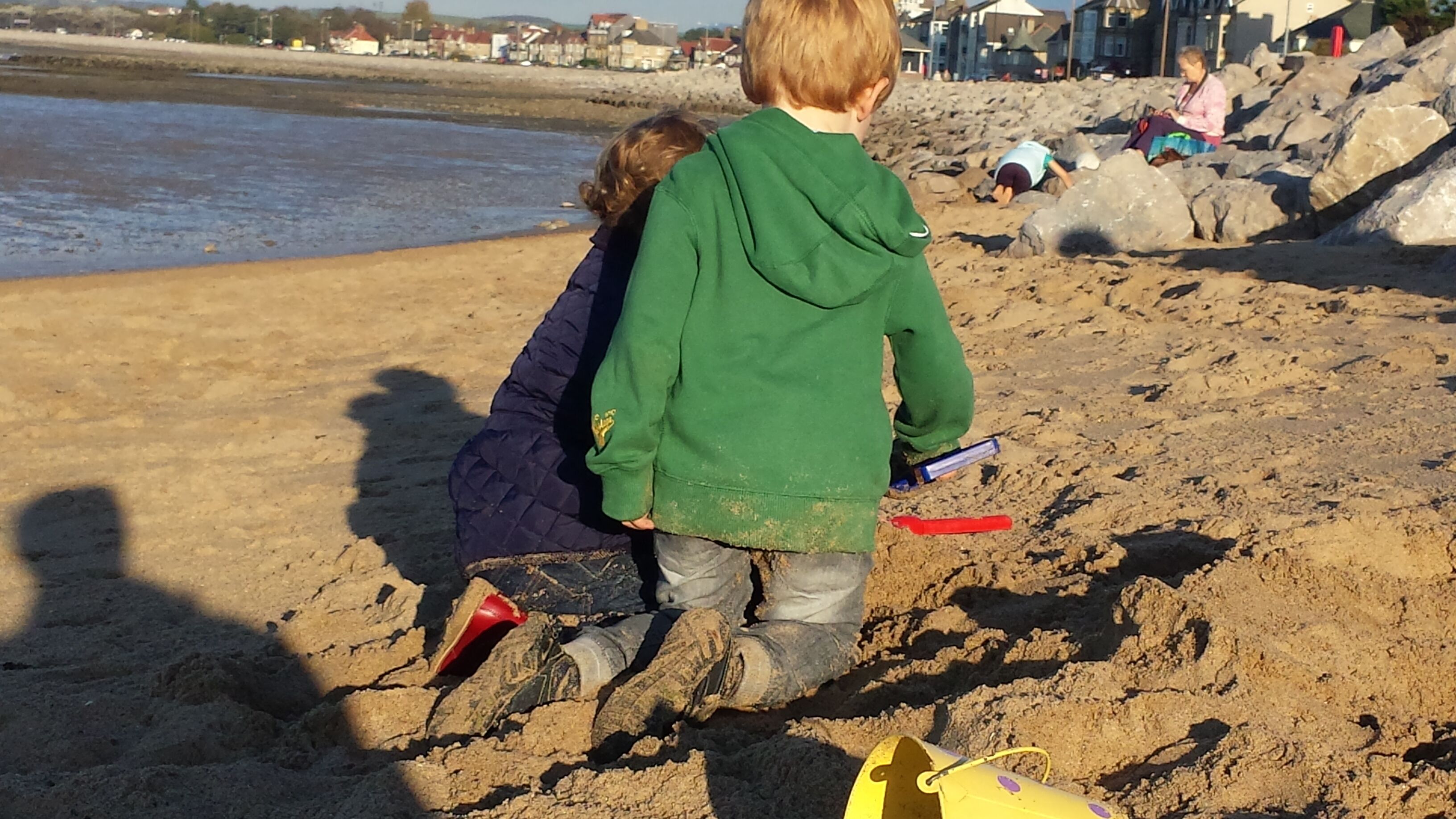 Two children playing on a sandy beach with rocks and a body of water in the background.