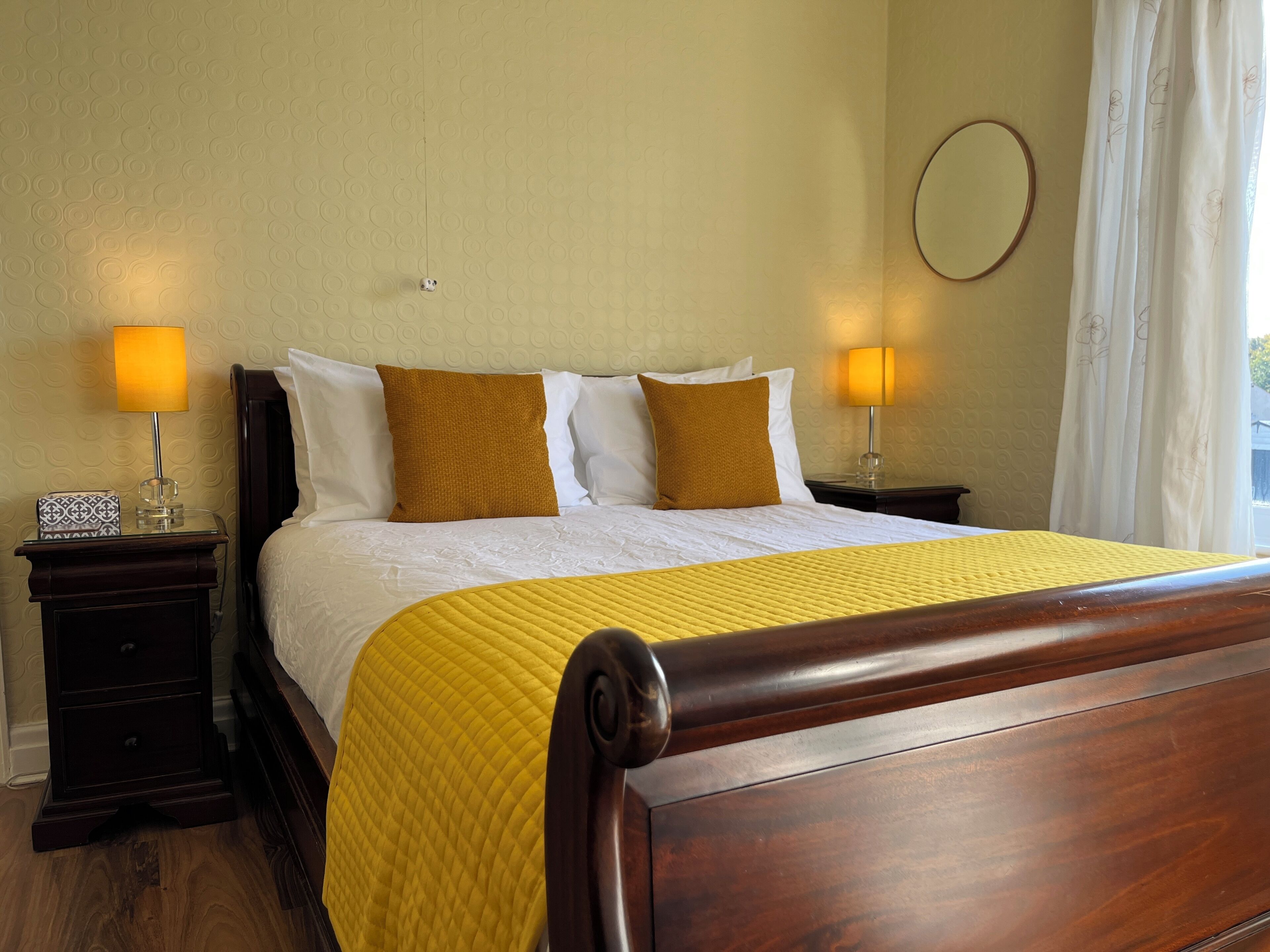 Una cama bien hecha con un cabecero de madera, dos mesitas de noche con lámparas, un espejo redondo y una ventana con cortinas.