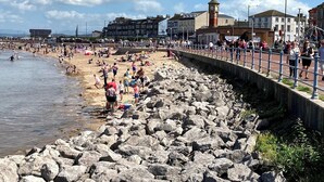 A rocky beach with people walking and sitting. There are buildings in the background and a clock tower. - 1 Lathom Avenue (Morecambe)