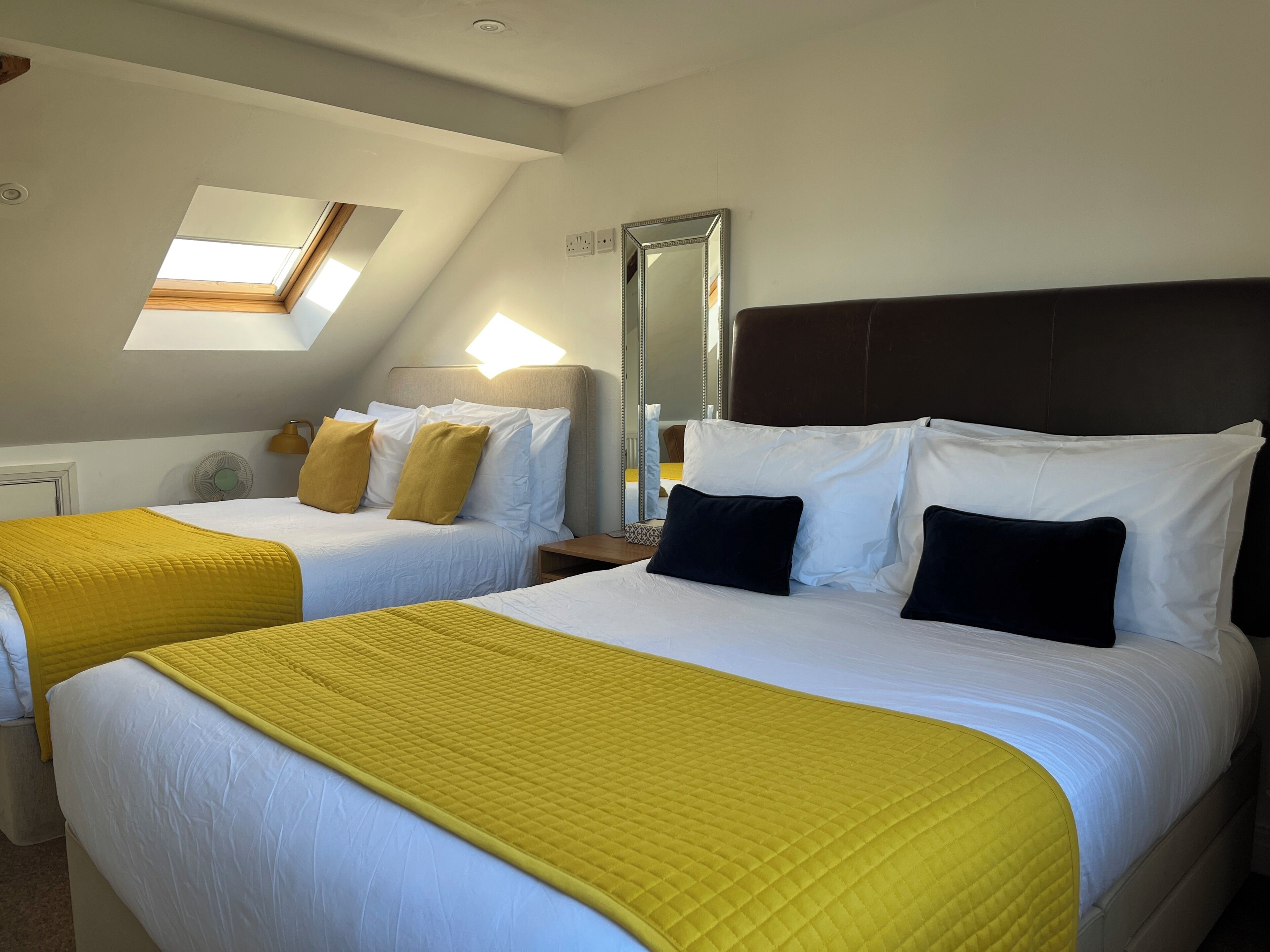 Habitación de hotel con dos camas, tragaluz, espejo y mesita de noche.