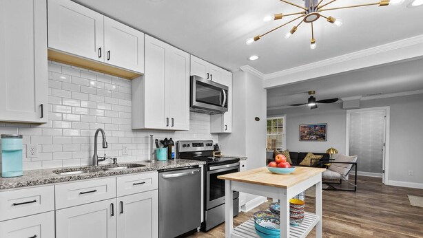 Una cucina moderna con mobili bianchi, un frigorifero in acciaio inossidabile e un forno a microonde. C'è una zona pranzo con tavolo e sedie, e un soggiorno con un divano e un quadro appeso al muro.