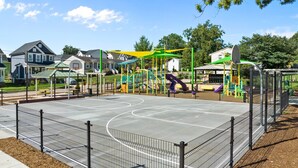 Un campo da basket recintato, circondato da giochi per bambini e case sullo sfondo.