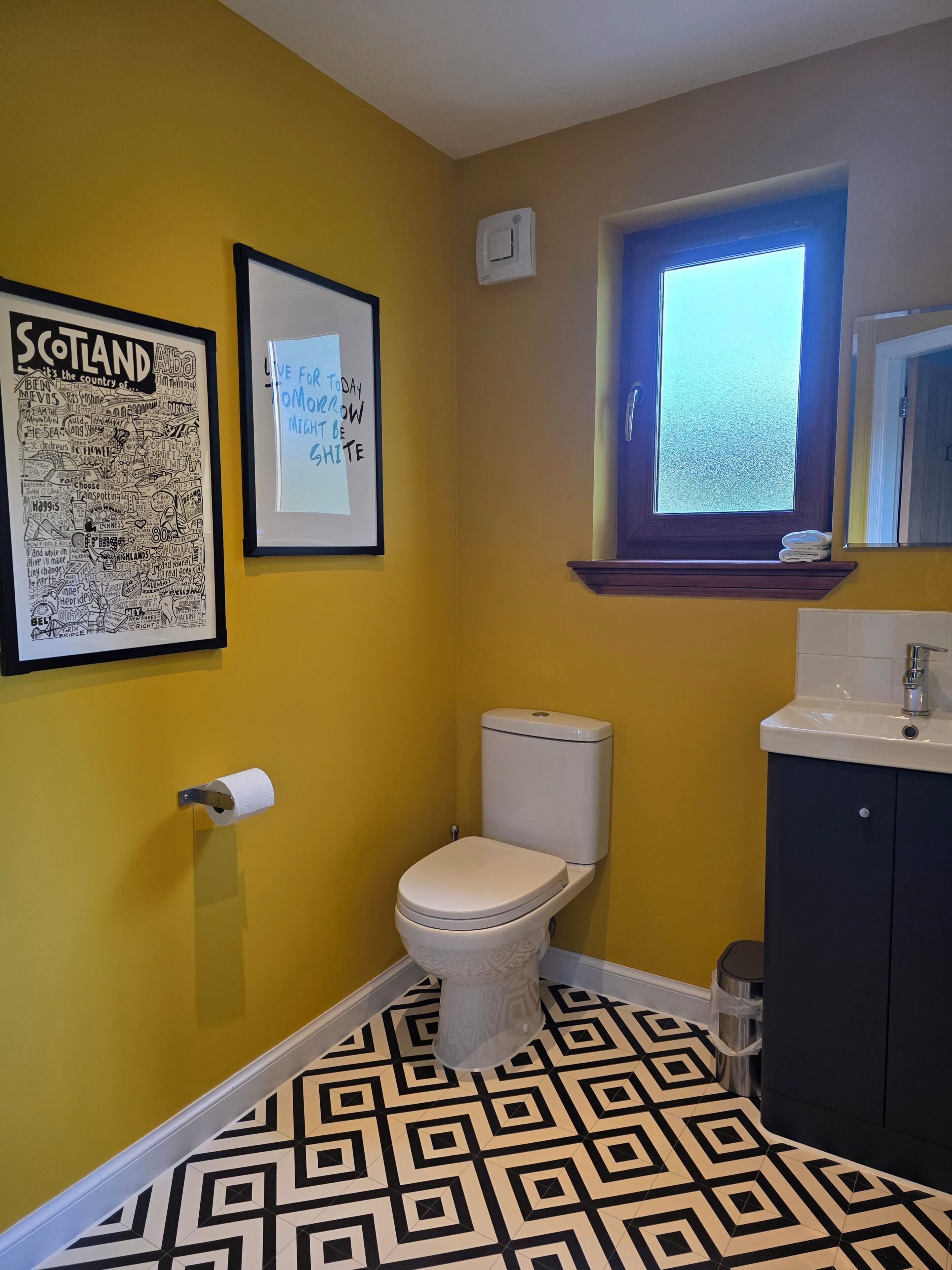 Une salle de bain avec un mur jaune, une carte de l’Écosse encadrée, un mot encadré, des toilettes, une fenêtre et un lavabo avec un miroir.