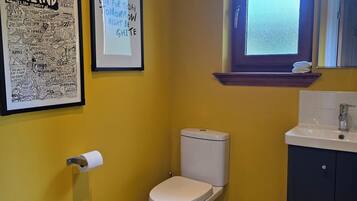 Une salle de bain avec un mur jaune, une carte de l’Écosse encadrée, un mot encadré, des toilettes, une fenêtre et un lavabo avec un miroir.