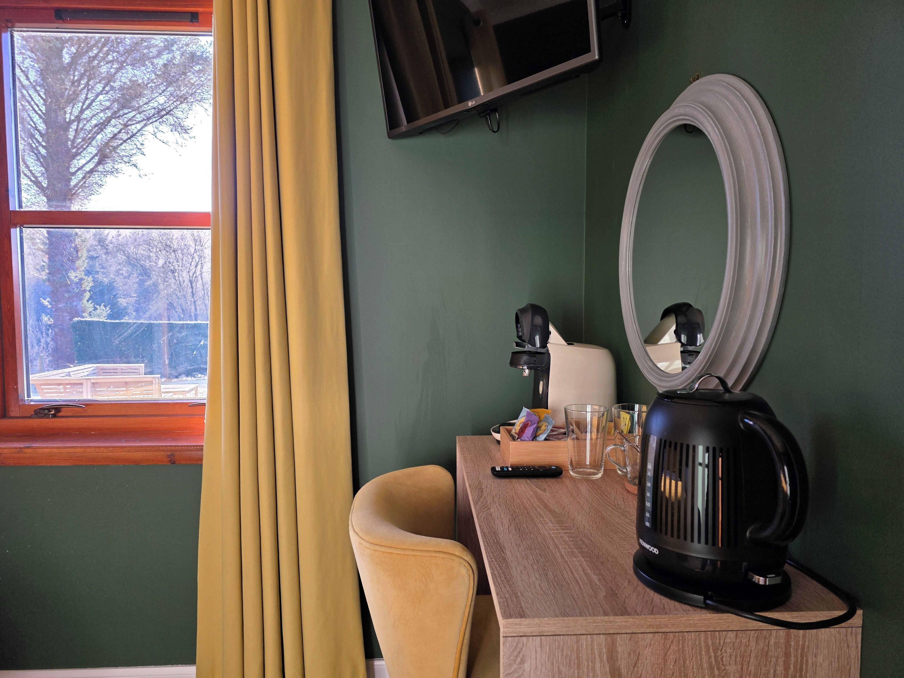A room with a wooden desk, a black coffee maker, a remote control, and a mirror on the wall.