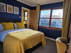 A bedroom with a bed, a chair, a window with a view, and artwork on the wall. - Mizpah Bed & Breakfast (Roybridge)