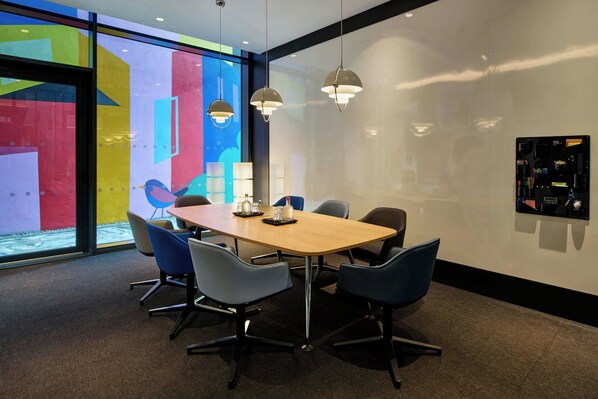 A modern meeting room with a wooden table, six chairs, pendant lights, and a large window with colorful artwork. - citizenM Dublin St. Patrick's (Dublin)