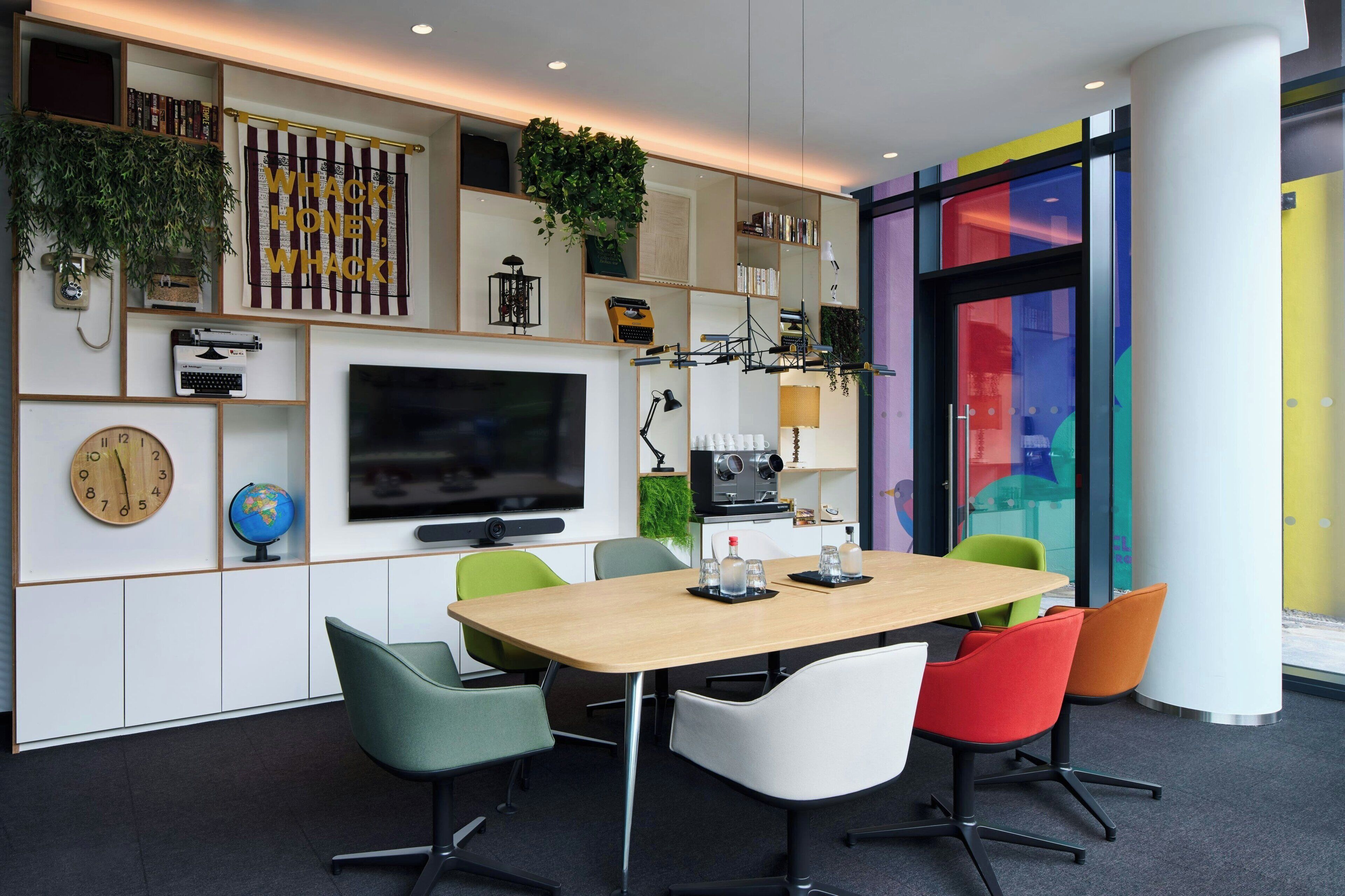 Una sala de reuniones moderna con una mesa de madera, sillas coloridas, un televisor de pantalla plana grande, estanterías y plantas decorativas.
