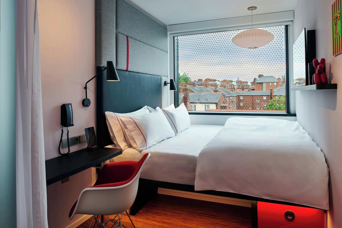 A modern hotel room with a large window, a bed, a desk, and a red chair.