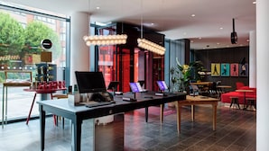 Una hall d'albergo moderna con una reception, schermi digitali e un'area lounge con sedie rosse.