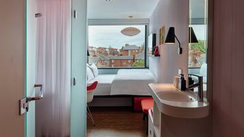 Une chambre moderne avec une grande fenêtre, un lit, un fauteuil rouge, une table de chevet blanche et une salle de bain avec un lavabo et un miroir.