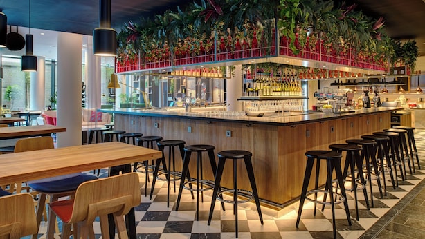 Un’area bar con pavimento a scacchi, tavoli in legno e sgabelli neri.
