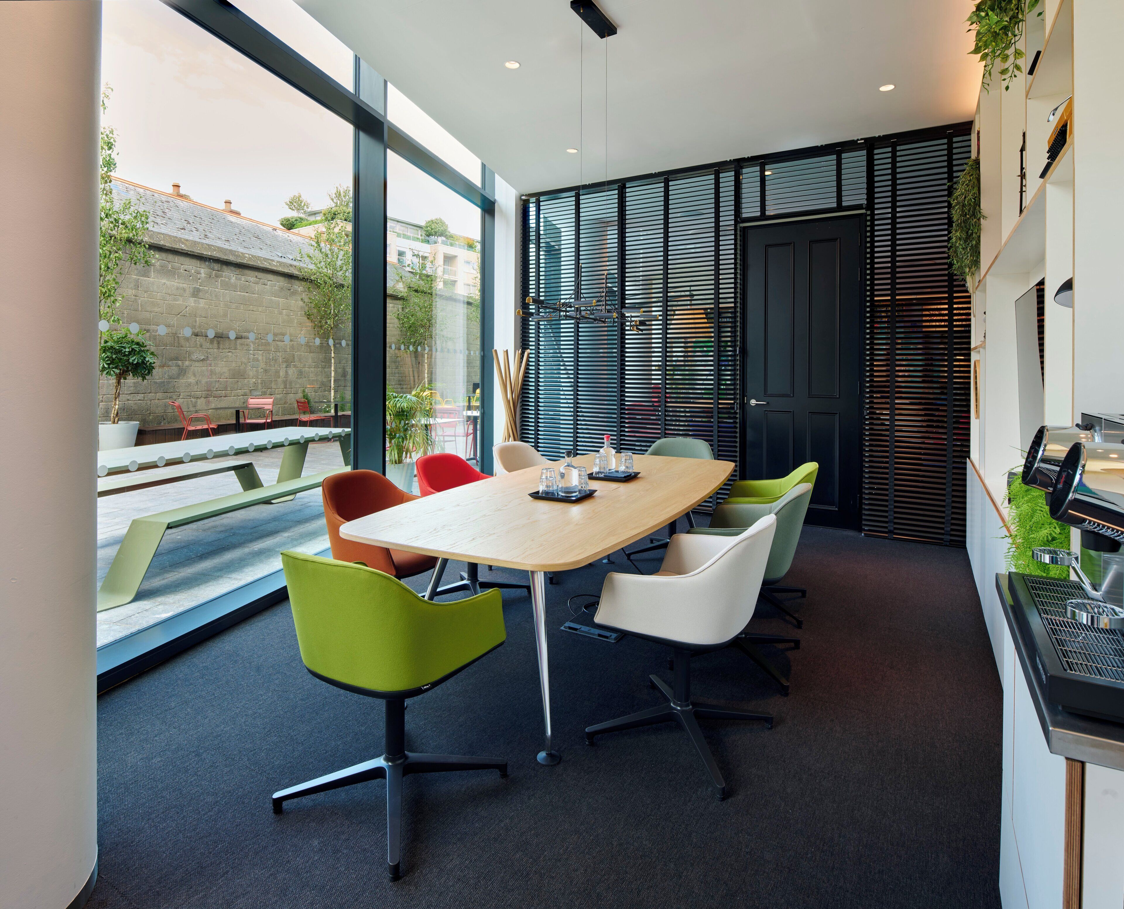 A modern meeting room with a large table, colorful chairs, and large windows offering a view of the outside.