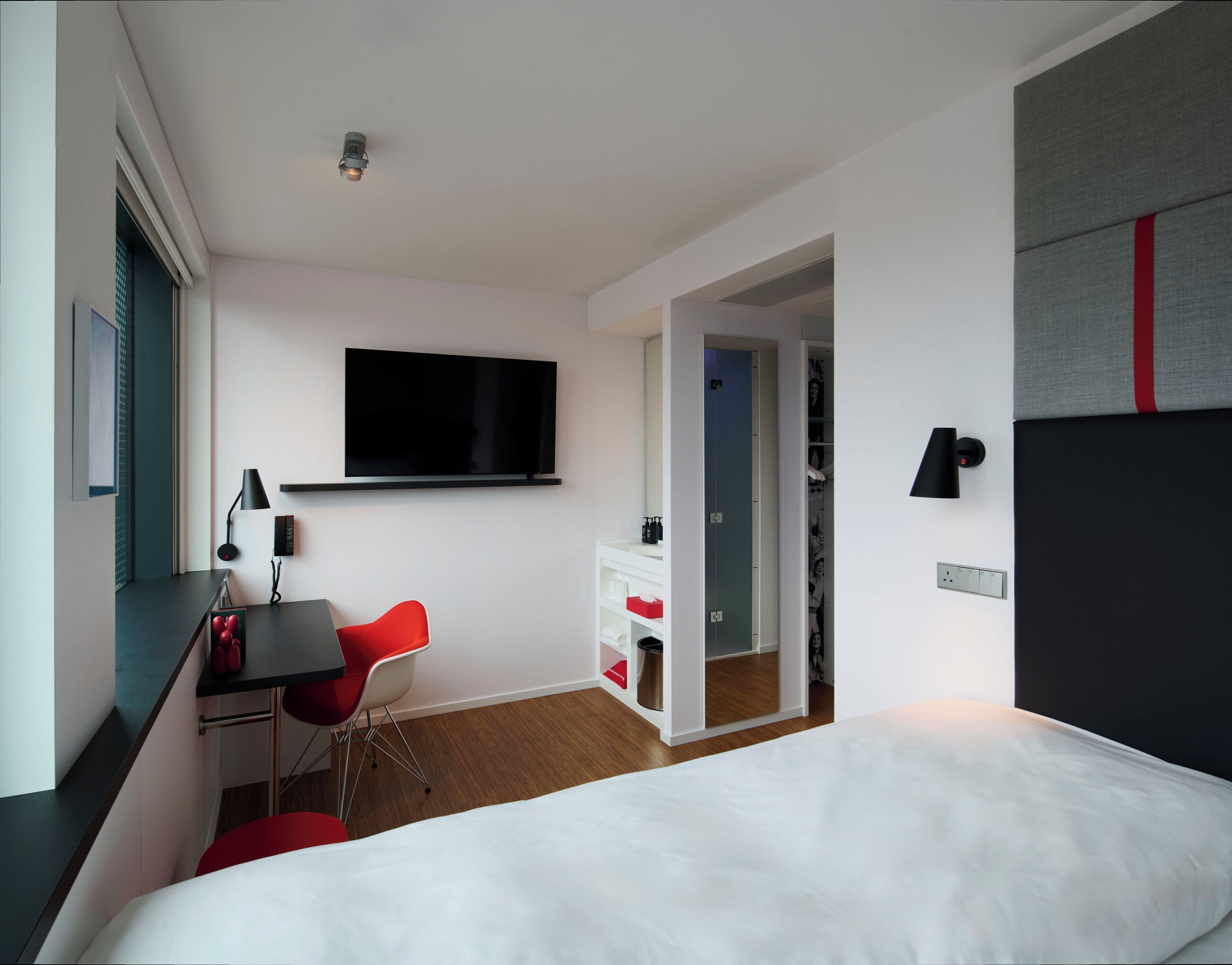 A modern hotel room with a bed, a desk with a chair, a flat-screen TV, a wardrobe, and a mirror.