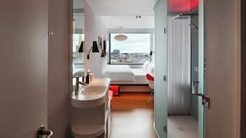 A modern hotel room with a large window, a bathroom with a sink and mirror, and a shower area.